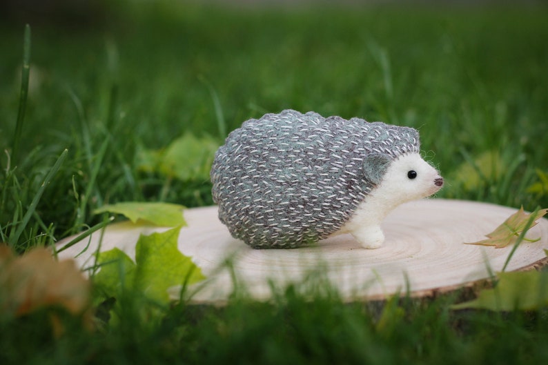 May include: A small, gray and white needle felted hedgehog figurine with a white belly and black eyes. The hedgehog is sitting on a wooden slice.