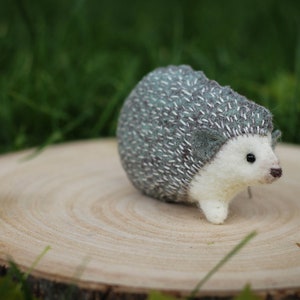 May include: A small, white and gray felt hedgehog with a stitched-on face and black eyes. The hedgehog is sitting on a wooden slice.