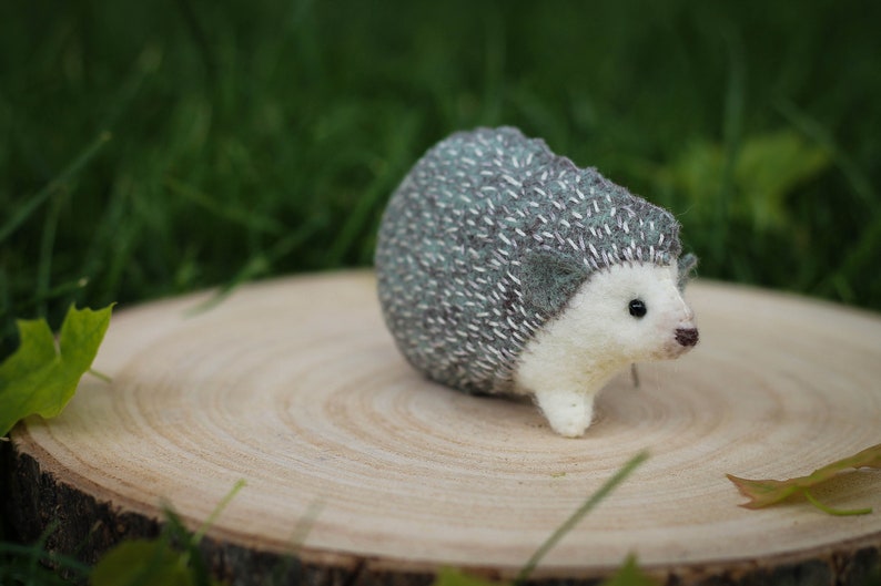 May include: A small, white and gray felt hedgehog with a stitched-on face and black eyes. The hedgehog is sitting on a wooden slice.