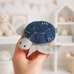 May include: A felt sea turtle plush toy featuring a dark blue shell with white stitched accents and a light gray head and flippers. The turtle has small black bead eyes and decorative white bead details on the shell segments.