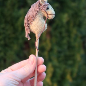 Puede incluir: Un peque&ntilde;o caballo de fieltro hecho a mano en un palo. El caballo es de color marr&oacute;n claro con una crin y cola rosadas. El palo es de madera y tiene una punta blanca.