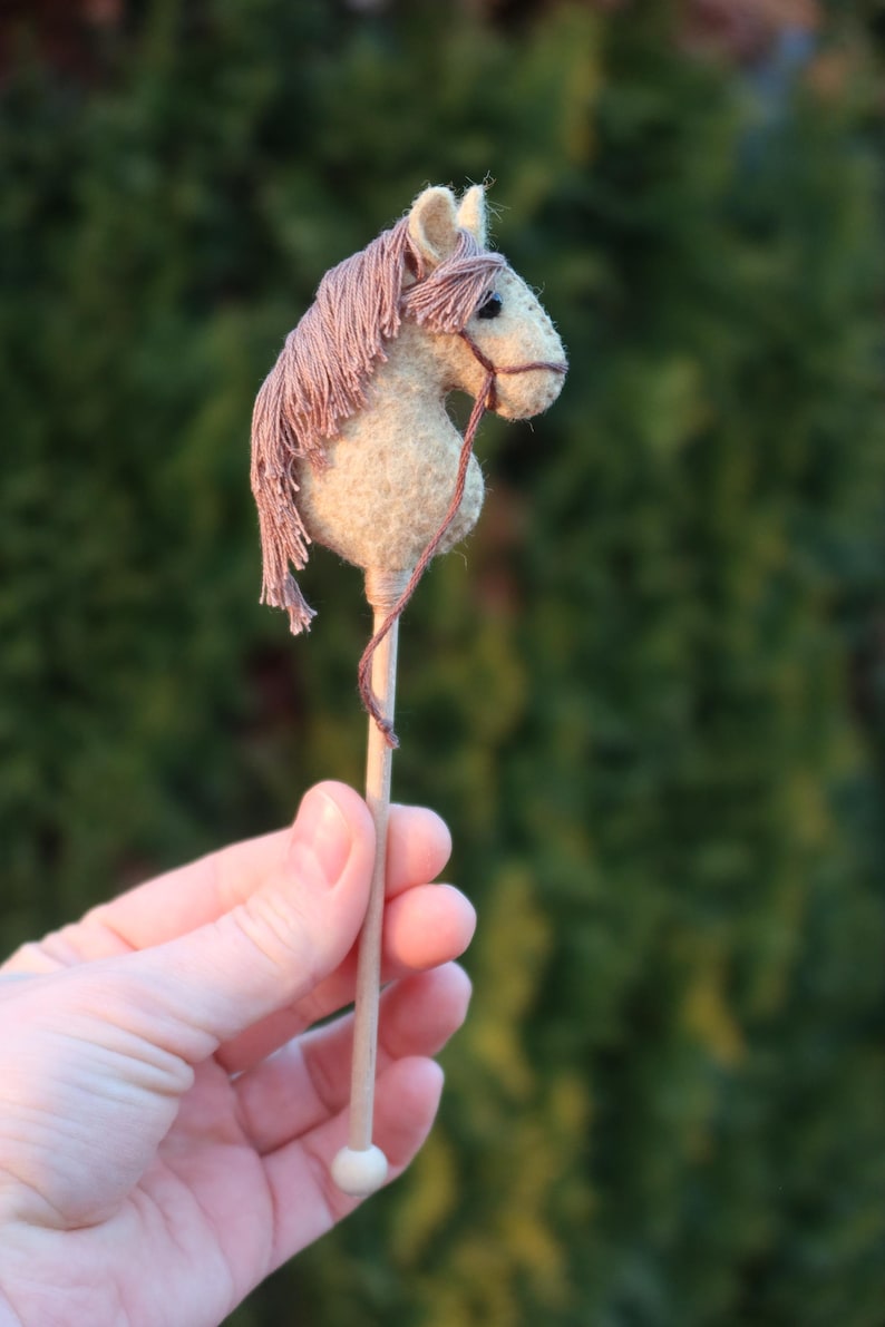 Puede incluir: Un peque&ntilde;o caballo de fieltro hecho a mano en un palo. El caballo es de color marr&oacute;n claro con una crin y cola rosadas. El palo es de madera y tiene una punta blanca.