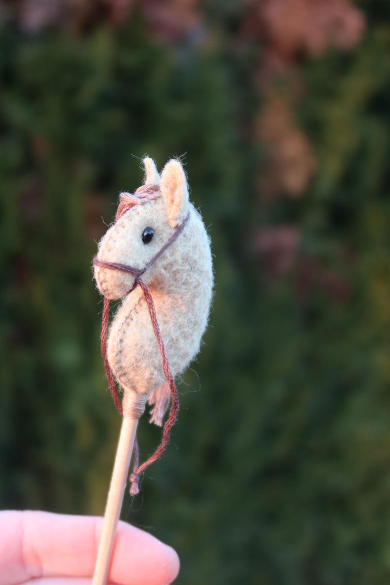 Puede incluir: Un peque&ntilde;o caballo de fieltro beige en un palo. El caballo tiene una brida marr&oacute;n y un ojo negro.