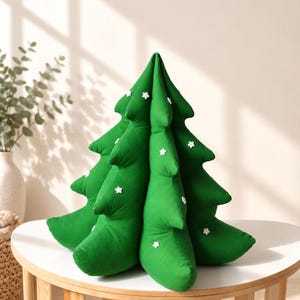 May include: A plush, cone-shaped Christmas tree pillow in a vibrant green fabric. The tree features multiple layered tiers of branches and is decorated with small, white, six-pointed snowflake-shaped embellishments scattered across the surface.