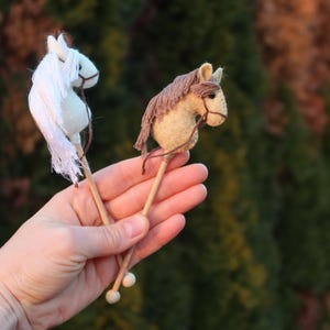 Puede incluir: Dos caballos de palo de madera, uno blanco y otro marr&oacute;n, se sostienen en la mano de una persona. Los caballos est&aacute;n hechos de fieltro y tienen una crin y una cola.