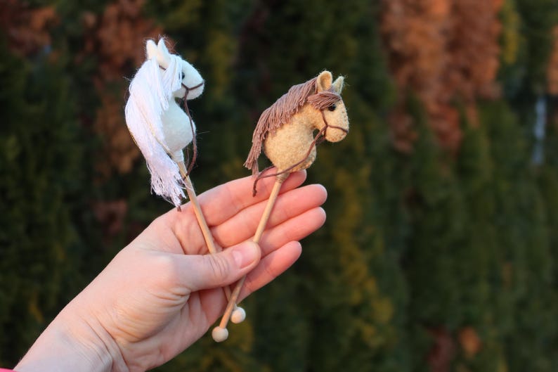Puede incluir: Dos caballos de palo de madera, uno blanco y otro marr&oacute;n, se sostienen en la mano de una persona. Los caballos est&aacute;n hechos de fieltro y tienen una crin y una cola.