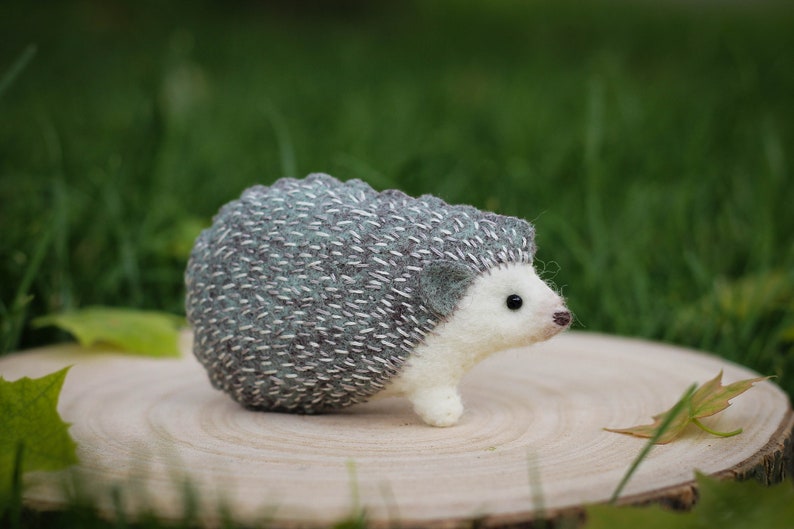 May include: A small, needle-felted hedgehog with a gray and white body. The hedgehog is sitting on a wooden slice.