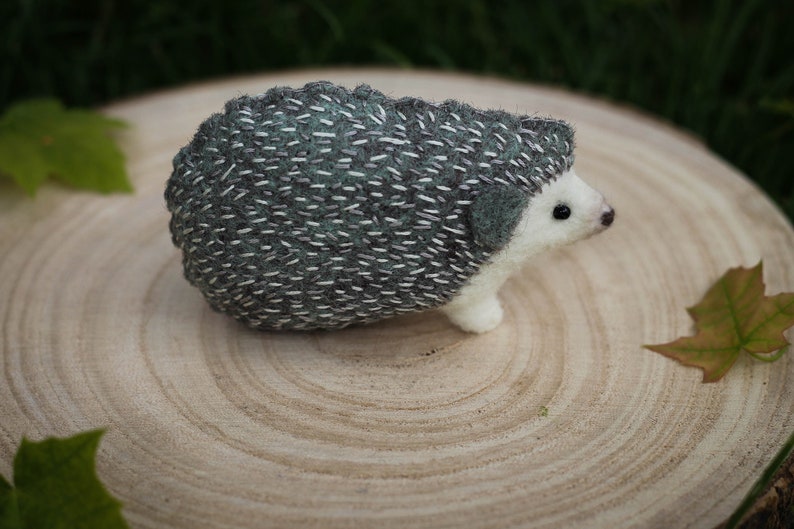 May include: A needle felted hedgehog with a gray and green spiky back and a white belly. The hedgehog is sitting on a wooden surface.