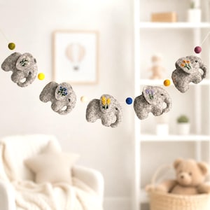 May include: A garland of gray felt elephants with embroidered floral details and colorful pom-poms. The elephants are strung together, creating a decorative banner. The garland is suitable for nursery or children's room decor.