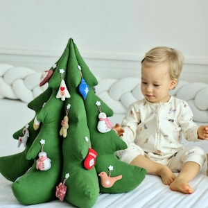 Pode incluir: Uma árvore de Natal de pelúcia verde com vários enfeites de feltro, incluindo um boneco de neve, um pássaro, um homem de gengibre e uma meia. Uma criança pequena está sentada em uma cama branca, olhando para a árvore.