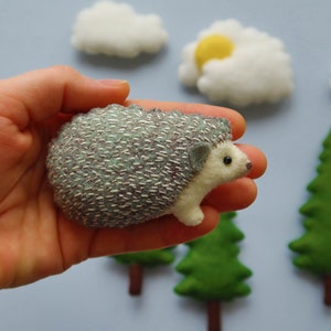 May include: A small, gray and white hedgehog with a silver embroidered design is held in a hand. The hedgehog is surrounded by felt clouds and trees.