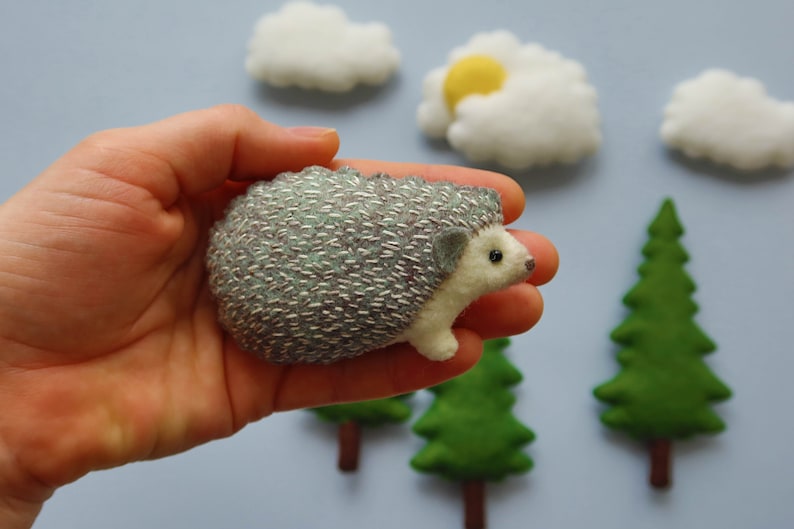 May include: A small, gray and white hedgehog with a silver embroidered design is held in a hand. The hedgehog is surrounded by felt clouds and trees.