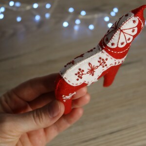 Może przedstawiać: Czerwona ozdoba konia Dala z filcu z białymi haftowanymi detalami. Koń jest trzymany w dłoni.