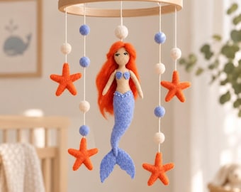 May include: A hanging mobile featuring a central felt mermaid with bright orange hair and a blue tail, surrounded by four orange felt starfish and alternating white and light blue felt spheres suspended from a circular wooden frame by white strings.