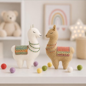 Op de afbeelding: Twee vilten lama-figuren, één wit en één beige, elk met gesloten ogen, geborduurde halskettingen en een gedessineerd zadeldek. De witte lama heeft een groen zadel, de beige lama een roze zadel, omringd door kleine vilten balletjes.