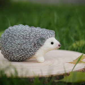May include: A small, hand-crafted hedgehog made of felt. The hedgehog is gray and white with a detailed, stitched design. It is sitting on a wooden surface.