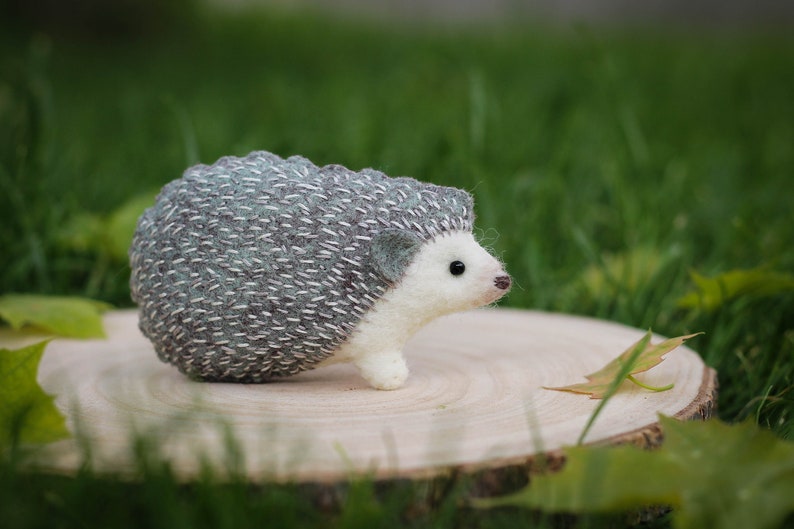 May include: A small, hand-crafted hedgehog made of felt. The hedgehog is gray and white with a detailed, stitched design. It is sitting on a wooden surface.
