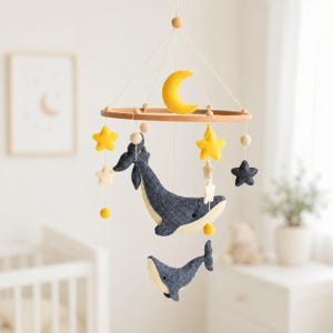 Könnte beinhalten: Ein Baby-Mobile mit einem Holzring, einem gelben Halbmond und Sternenverzierungen. Das Mobile enthält zwei dunkelblaue Wale, gelbe Sterne und kleine weiße Sterne, die eine skurrile Kinderzimmerdekoration schaffen.