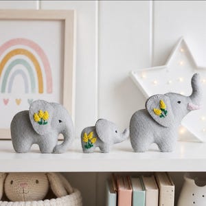 May include: Three grey felt elephant figurines of varying sizes, each featuring embroidered yellow flowers on the side. The elephants have stitched black eyes and light pink accents on their trunks, arranged in a row on a white shelf.