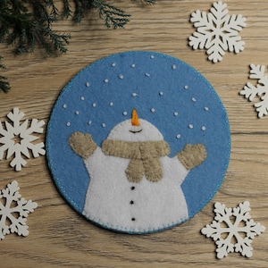 May include: A felt snowman ornament with a blue background and white snowflakes. The snowman is wearing a brown scarf and has a carrot nose.