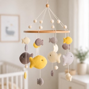 May include: A baby mobile featuring felt fish in shades of yellow, gray, and cream, suspended from a wooden hoop. The mobile includes small felt balls and is designed for a nursery.