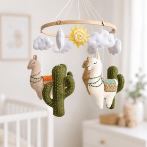 Puede incluir: Un móvil colgante con figuras de fieltro de dos llamas, dos cactus verdes, un sol amarillo y nubes blancas suspendidas de un marco circular de madera. Las llamas tienen mantas decorativas y detalles cosidos, mientras que las nubes y el sol son suaves.