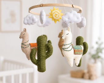 May include: A hanging mobile featuring felt figures of two llamas, two green cacti, a yellow sun, and white clouds suspended from a circular wooden frame. The llamas have decorative blankets and stitched details, while the clouds and sun are soft.