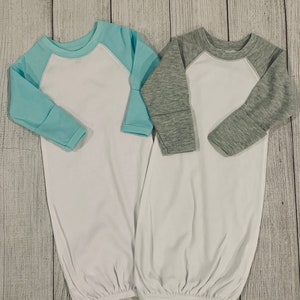 May include: Two white baby gowns with long sleeves. One gown has a light blue baseball-style sleeve and the other has a gray baseball-style sleeve.