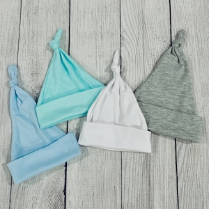 May include: Four baby hats in different colors and styles. The hats are made of soft, stretchy fabric and are perfect for newborns. The hats are laid out on a white wooden surface.