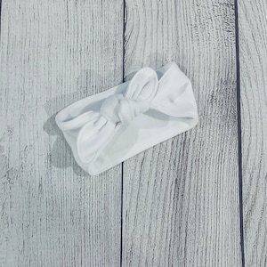 May include: A white knotted headband made of soft fabric. The headband is tied in a bow shape.