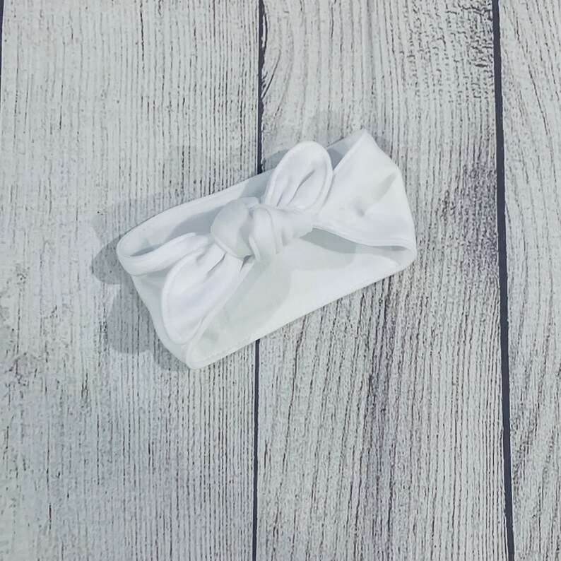 May include: A white knotted headband made of soft fabric. The headband is tied in a bow shape.