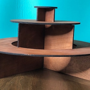 Pode incluir: Um suporte de exibição de madeira de três níveis com forma circular. O suporte é feito de madeira marrom e tem um acabamento natural. Cada nível é ligeiramente menor que o de baixo, criando um efeito em camadas.