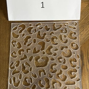May include: A clear plastic stencil with a leopard print design. The stencil is square and has a number "1" written on a white card behind it.