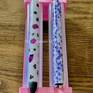 May include: Two pens in a pink holder. One pen is white with black and pink spots. The other pen is blue with a white floral pattern. The pens are in a pink holder with a rectangular base.