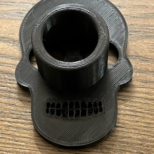 May include: Black 3D printed skull-shaped phone stand with a round hole in the top for holding a phone.