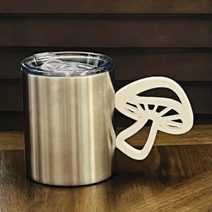 May include: A stainless steel travel mug with a white mushroom-shaped handle. The mug has a clear lid.
