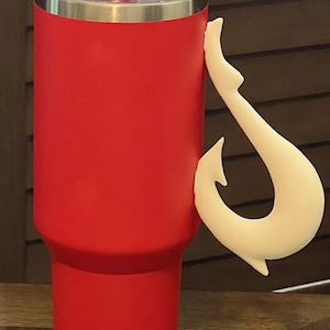 May include: A red insulated tumbler with a clear lid and a cream-colored hook-shaped handle. The tumbler is cylindrical and appears to be made of stainless steel. The handle is attached to the side of the tumbler.