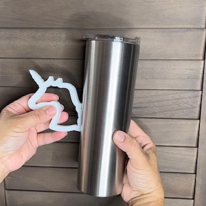 May include: A stainless steel tumbler with a clear lid is held with a white, decorative handle. The tumbler is cylindrical and has a silver finish. The handle is in the shape of a unicorn.