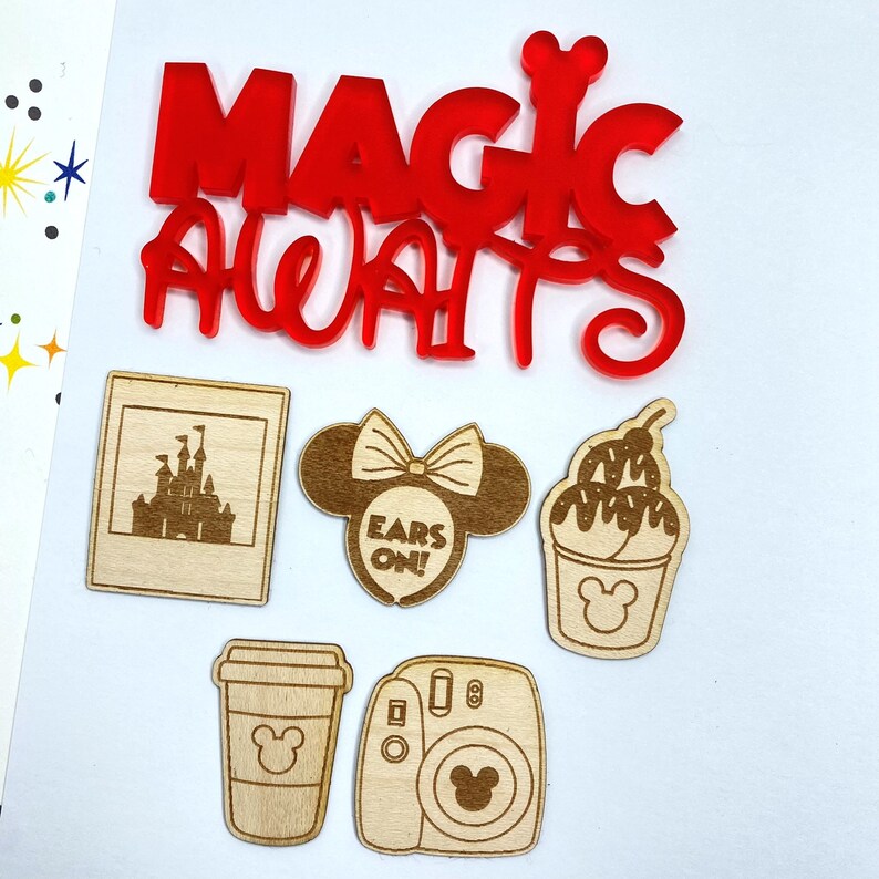 Op de afbeelding: Een set van zes houten lasergesneden ornamenten met een kasteel, Mickey Mouse-oren, een beker ijs, een koffiebeker, een camera en een rood acryl bord met de tekst "Magic Awaits".