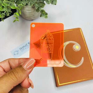May include: Orange acrylic stencil with a white outline of a pumpkin spice latte. The stencil is translucent and has a hole in the top corner.