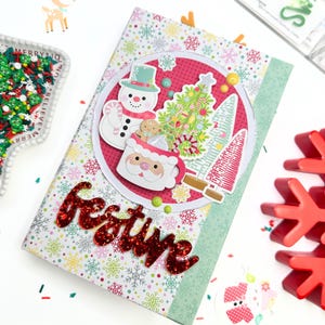 May include: A festive Christmas-themed journal with a red glitter word "Festive" on the cover. The journal features a snowman, Christmas tree, and Santa Claus mug design. The background has a snowflake pattern.