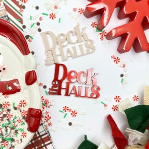 Puede incluir: Dos letreros de madera con el texto "Deck the Halls" en letras blancas y rojas. Los letreros están sobre un fondo blanco con chispas rojas y blancas.