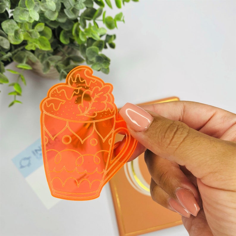 May include: Orange acrylic sticker of a coffee mug with whipped cream and a leaf design. The mug has a decorative pattern.
