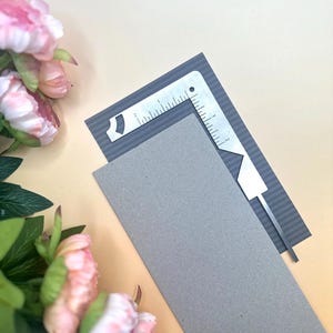 Peut inclure: Une règle d'angle en métal en forme de L avec des graduations jusqu'à 5 cm, posée sur une carte rectangulaire gris foncé rayée et une carte grise unie, avec des pivoines roses visibles dans le coin du cadre.