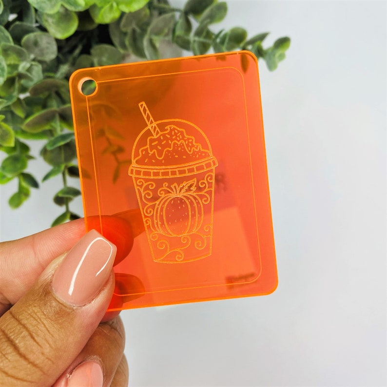 May include: Orange acrylic keychain with a white outline of a pumpkin spice latte. The latte has a straw and whipped cream with sprinkles.