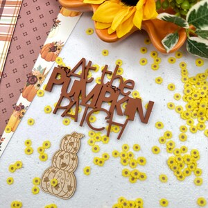 May include: Wooden sign with the text "At the Pumpkin Patch" in brown letters. A wooden pumpkin stack with fall leaves is in the lower left corner. The background is white with yellow flower sprinkles.