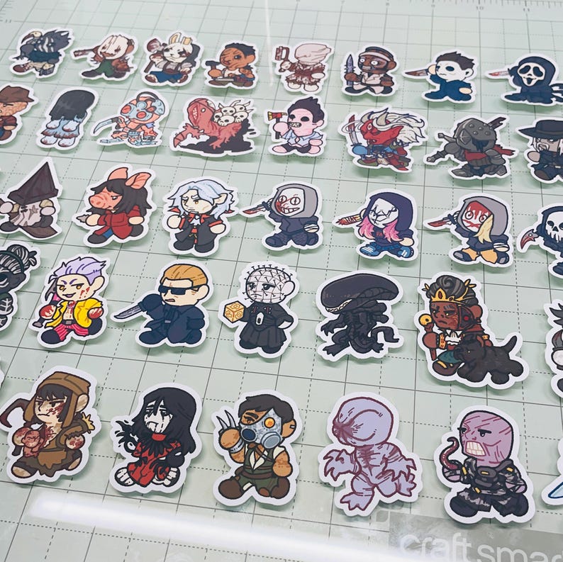 May include: A collection of small, die-cut vinyl stickers featuring chibi-style illustrations of various horror movie characters and creatures, arranged in a grid pattern on a light green cutting mat with a white grid line overlay.