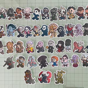 May include: A collection of forty-five individual die-cut stickers featuring chibi-style illustrations of various horror movie and video game characters, including figures with masks, blades, and supernatural features, arranged on a green grid mat.