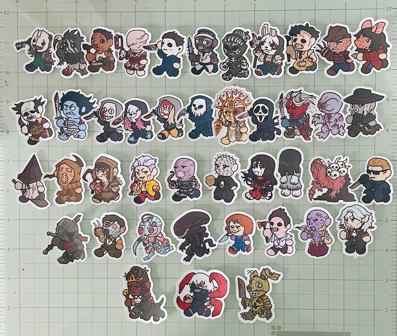 May include: A collection of forty-five individual die-cut stickers featuring chibi-style illustrations of various horror movie and video game characters, including figures with masks, blades, and supernatural features, arranged on a green grid mat.