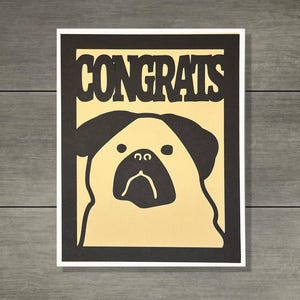 以下が含まれることがあります： 黒い縁取りと太字の「CONGRATS」の文字が入ったグリーティングカード。クリーム色の顔と黒い耳を持つパグ犬の様式化されたイラストが特徴です。カードは灰色の木製の背景に配置されています。